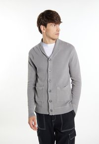 Šedý strukturální cardigan s šálovým límcem, šesti předními knoflíky a dvěma předními kapsami. Nosený přes bílou košili, v kombinaci s tmavými kalhotami.