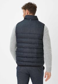 Dunkelblauer Daunenweste mit hohem Kragen, mit gestepptem Muster und Reißverschluss. Getragen über einem hellgrauen Pullover.