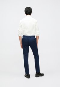 Balti ilgomis rankovėmis marškiniai, tamsiai mėlynos kelnės su kišenių detalėmis, sklandus audinys, siauras prigludimas, derinamos su juodais batais, matoma iš nugaros.