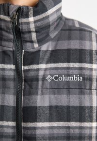 Pelēkā šķērsota jaka ar rāvējslēdzēju un augstu apkakli, ar melnbaltiem rakstiem un nelielu Columbia logotipu kreisajā krūtī.