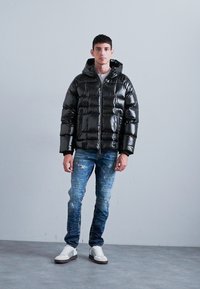 Giubbotto a vento nero con finitura lucida, chiusura con zip e cappuccio, abbinato a jeans blu con effetto consumato e sneaker bianche. L'ambiente è di un grigio neutro.