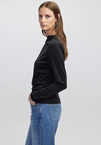 Langärmliges schwarzes Oberteil mit hohem Kragen und seitlichen Raffung, kombiniert mit blauen Jeans aus Denim, die seitliche Nietenakzente aufweisen.