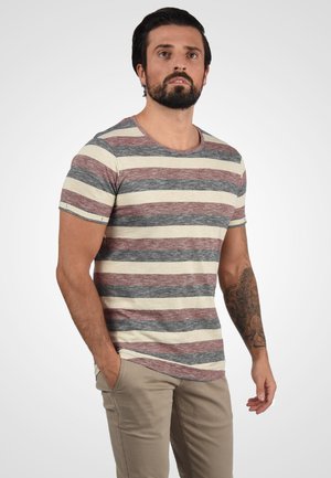 Mand med skæg og tatovering iført en kortærmet, stribet t-shirt og beige bukser, stående mod en ensartet lys baggrund.