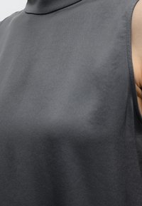 Gros plan d'une personne portant un haut sans manches à col roulé gris foncé avec une texture de tissu visible et une partie du bras montrée.