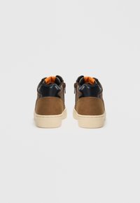 Încălțăminte înaltă din suede maro cu accente din piele neagră, talpă din cauciuc alb și căptușeală portocalie. Dispun de două fermoare laterale pentru a fi ușor de purtat.