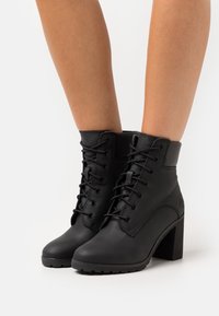 Černé kožené kotníkové boty s tkaničkami, pevnou patou a texturovanou podrážkou. Mají hladký svršek s jemným detaily švů.