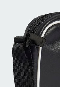 Kôš na syntetickej taške v čiernej farbe s textúrovaným povrchom, bielym akcentovým pruhom a zipsom so čiernym ťahadlom. Nastaviteľný tkáčsky popruh.