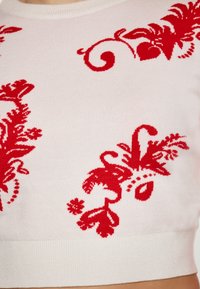 Motivi floreali rossi su un pullover corto bianco, con orlo a costine e una consistenza morbida, che evidenzia dettagli di design intricati.
