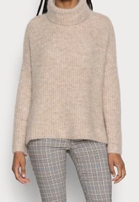Maglione beige morbido con collo a lupetto e texture a coste. Fit morbido con maniche lunghe, abbinato a pantaloni a quadri con tonalità marroni e nere.
