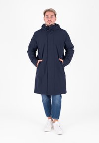 Cappotto impermeabile blu navy con cappuccio, chiusura con zip e tasche laterali, abbinato a jeans azzurri e sneakers bianche.