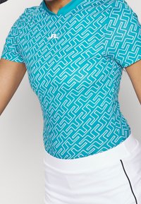 Turquoise sportshirt met een wit zigzagpatroon. Korte mouwen, ronde halslijn en een getailleerd ontwerp. Draagt bij een witte onderkant.