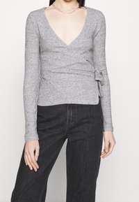 Grå, figurnära långärmad topp med djup V-ringning och omslagdesign. Texturerad tyg med en sidoknytdetalj. Kombinerad med svarta högmidjade jeans.