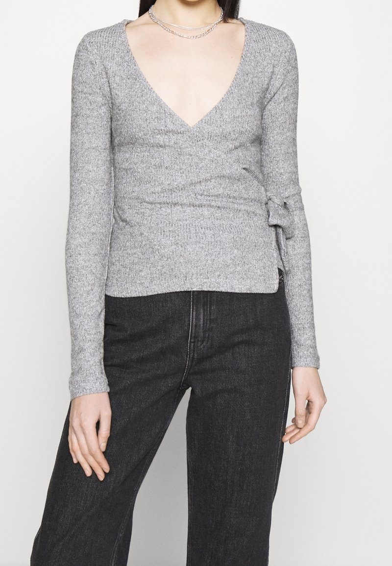 Grå, figurnära långärmad topp med djup V-ringning och omslagdesign. Texturerad tyg med en sidoknytdetalj. Kombinerad med svarta högmidjade jeans.