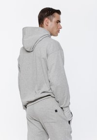 Szara bluza z kapturem, wykonana z miękkiego materiału, z dużym kapturem, luźnym krojem i widocznymi szwami. Połączona z pasującymi spodniami dresowymi z kieszeniami.