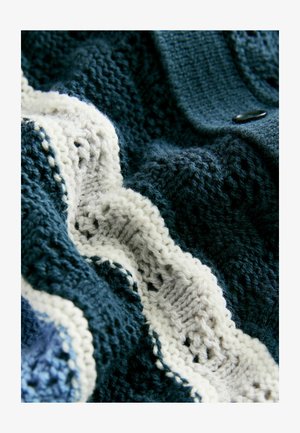 Adīta tekstila ar tumši ziliem un baltajiem svītrainiem, demonstrējot teksturālus rakstus ar mīkstu, pūkainu izskatu un pogu akcentu.