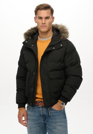 Mężczyzna w czarnej puchowej kurtce z futrzanym kapturem, pomarańczowym swetrze, niebieskich jeansach, brązowym pasku i zegarku na nadgarstku, stojący na białym tle.