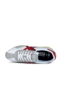 Zapatilla blanca con detalles en rojo y gris. Presenta una puntera de ante gris texturizado, cordones blancos y una plantilla negra con detalles de talla.