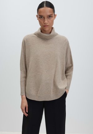 Femme aux cheveux foncés portant un pull à col roulé beige et un pantalon noir, se tenant devant un fond uni gris clair.