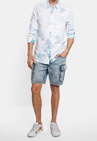Camicia bianca a maniche lunghe con motivi floreali blu, abbinata a pantaloni cargo azzurri chiari e sneaker multicolori. Look casual, vestibilità comoda.