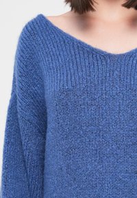 Maglione blu lavorato a maglia con una ampia scollatura a V, texture a coste e maniche ampie. Il tessuto appare morbido e peloso.