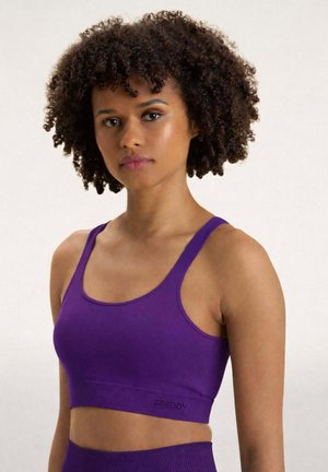 Donna con capelli ricci che indossa un reggiseno sportivo viola aderente e leggings a vita alta abbinati, in piedi davanti a uno sfondo semplice.