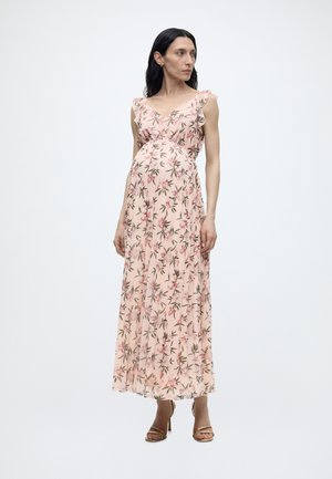 Έγκυος γυναίκα που φοράει αμάνικο ροζ πολύχρωμο μακρύ φόρεμα και μπεζ ψηλοτάκουνα σανδάλια, στέκεται και κοιτάζει προς τα δεξιά.