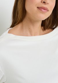 Top in tessuto morbido bianco con scollo a barca e vestibilità comoda, caratterizzato da una texture liscia e dettagli di cucitura minima lungo i bordi.