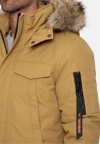 Zandkleurig geïsoleerd jack met een capuchon van nepbont, een ritszak op de mouw, klepzakken aan de voorkant en stevige ritsen.