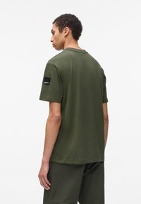 T-shirt verde măsliniu cu mânecă scurtă din bumbac ușor, cu guler la baza gâtului, având o etichetă neagră pe mâneca stângă. Textură fină.