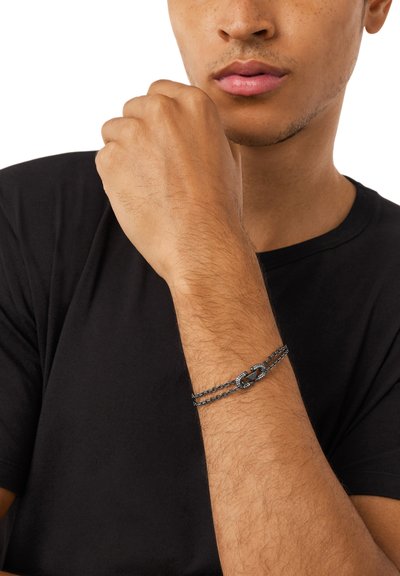 Homme portant un bracelet chaîne noir au poignet, bras levé près du visage, habillé d'une chemise noire, fond neutre.