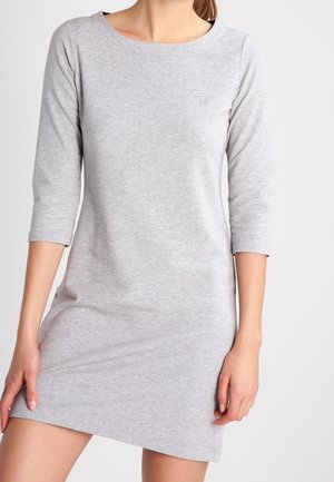 Kvinde iført en ensfarvet lysegrå knælængde kjole med trekvart ærmer, stående mod en hvid baggrund.