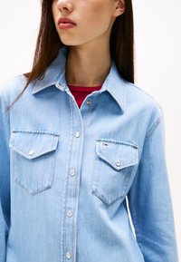 Svijetloplava traper košulja s oštrim kragnom, dva džepa na prsima, srebrnim dugmadi i detaljima šavova. Nosiva preko crvene bluze.