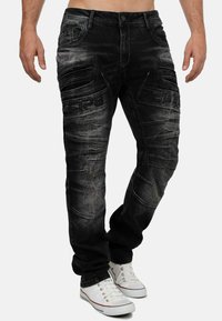 Pantalons cargo en denim noir avec une finition délavée, comportant plusieurs poches, des détails cousus et des panneaux texturés, associés à des baskets blanches.