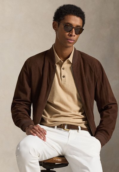 Giacca bomber marrone sopra un polo beige con due bottoni. Pantaloni bianchi e cintura marrone. Posizione seduta con le mani appoggiate su uno sgabello di legno.