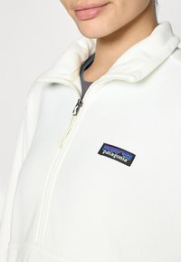 Sudadera de forro polar blanca con alto cuello y cremallera de un cuarto. Presenta un parche del logo negro de Patagonia en el pecho. Textura suave.