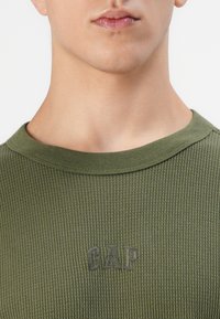 Nærbilde av en person som har på seg en olivengrønn teksturert skjorte med rund hals og mørk brodert "GAP"-logo på brystet.