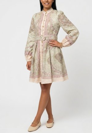 Kobieta nosząca sukienkę sięgającą do kolan, z długim rękawem i paskiem, z wzorem paisley w delikatnych beżowych, mauve i zielonych odcieniach, w połączeniu z beżowymi balerinami.