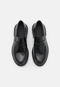 Mocassins en cuir noir avec une finition brillante, dotés d'une semelle plate et d'un design classique à fente pour pièce sur le dessus, avec un branding minimal.