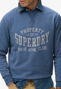 Sudadera azul con cuello redondo, que presenta un logo gris impreso "PROPERTY OF SUPERDRY STATE ATHL CLUB" en el frente y puños acanalados.