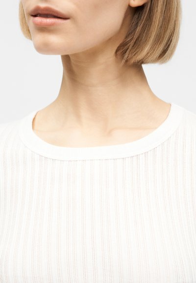 Κοντινό πλάνο στο κάτω μέρος του προσώπου και τον λαιμό μιας γυναίκας με κοντά ξανθά μαλλιά, που φορά λευκή ριμπ μπλούζα με στρογγυλή λαιμόκοψη.