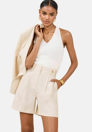 Pantaloni corti in lino beige con pieghe e tasche laterali, abbinati a una canotta bianca aderente a costine e a un blazer leggero. Bracciale e orecchini dorati.