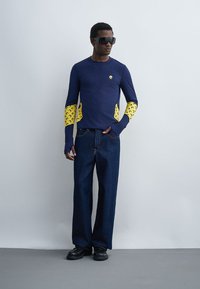 Mees kannab mereväe sinist pikade varrukatega särki kollaste poolkuu detailidega varrukatel ja külgedel, tumedaid laia säärega teksaseid, musti kingi ja suuri musta päikeseprille.