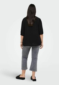 Maglietta nera a maniche corte con spalle texturizzate, abbinata a jeans grigi a vita alta e scarpe slip-on nere. La modella è di spalle rispetto all'osservatore.