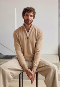 Beiger Strickpullover mit Reißverschluss und weicher Textur, kombiniert mit hellfarbenen Hosen. Das Modell sitzt auf einem Metallhocker vor einem neutralen Hintergrund.