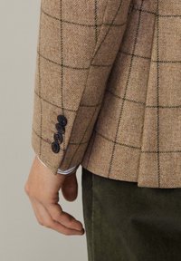 Mano di un uomo appoggiata al fianco, che indossa un blazer a quadri beige con quattro bottoni neri sui polsini e pantaloni di velluto a coste verde scuro.