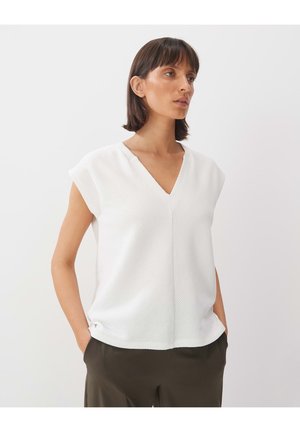 Weiße V-Ausschnitt-Bluse mit kurzen Ärmeln, strukturiertem Muster und lockerer Passform. Kombiniert mit dunkelgrünen Hosen, die ein schlichtes Design betonen.