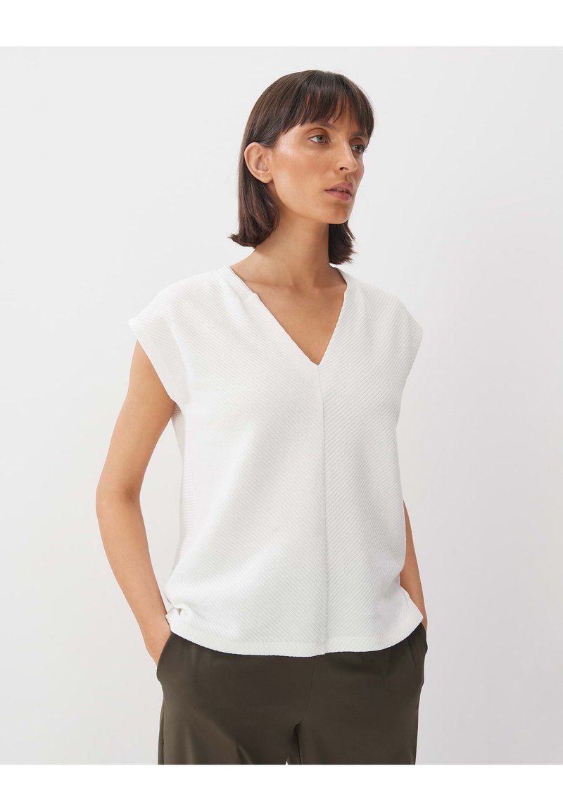 Weiße V-Ausschnitt-Bluse mit kurzen Ärmeln, strukturiertem Muster und lockerer Passform. Kombiniert mit dunkelgrünen Hosen, die ein schlichtes Design betonen.