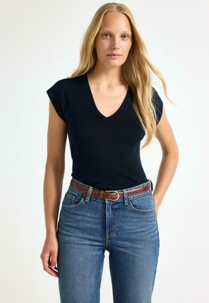 Ung kvinde med langt blondt hår iført sort V-hals top, blå jeans og brunt bælte, stående mod en ensartet lys baggrund.