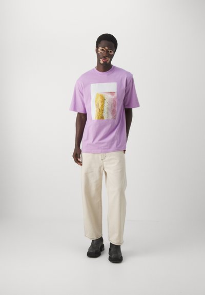 Lila kortekorte t-shirt met een vierkant grafisch ontwerp van gelaagde dessert, gedragen met off-white wide-leg broek en zwarte enkellaarsjes.