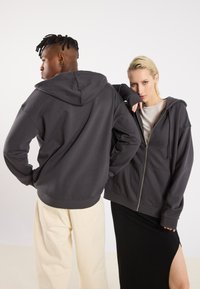 Siva zip-up dukserica od mekog materijala, koja ima kapuljaču, opušteni kroj i rebraste manžete, u kombinaciji sa svijetlim hlačama i crnom suknjom.